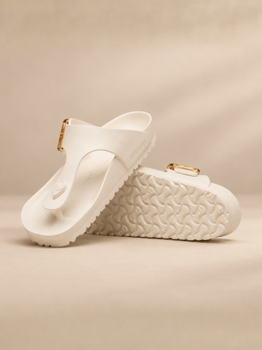 A pair of white thong sandals with textured soles and small gold-tone buckle details, displayed on a warm beige background.