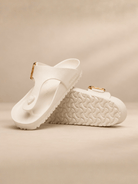 A pair of white thong sandals with textured soles and small gold-tone buckle details, displayed on a warm beige background.