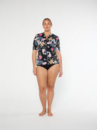 Woman wearing a floral short-sleeve shirt and black swimsuit bottom on a white background