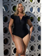 Woman wearing a black swimsuit standing against a geometric-patterned wall.