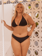 Woman wearing a black bikini with hipster pant standing against a decorative wall.