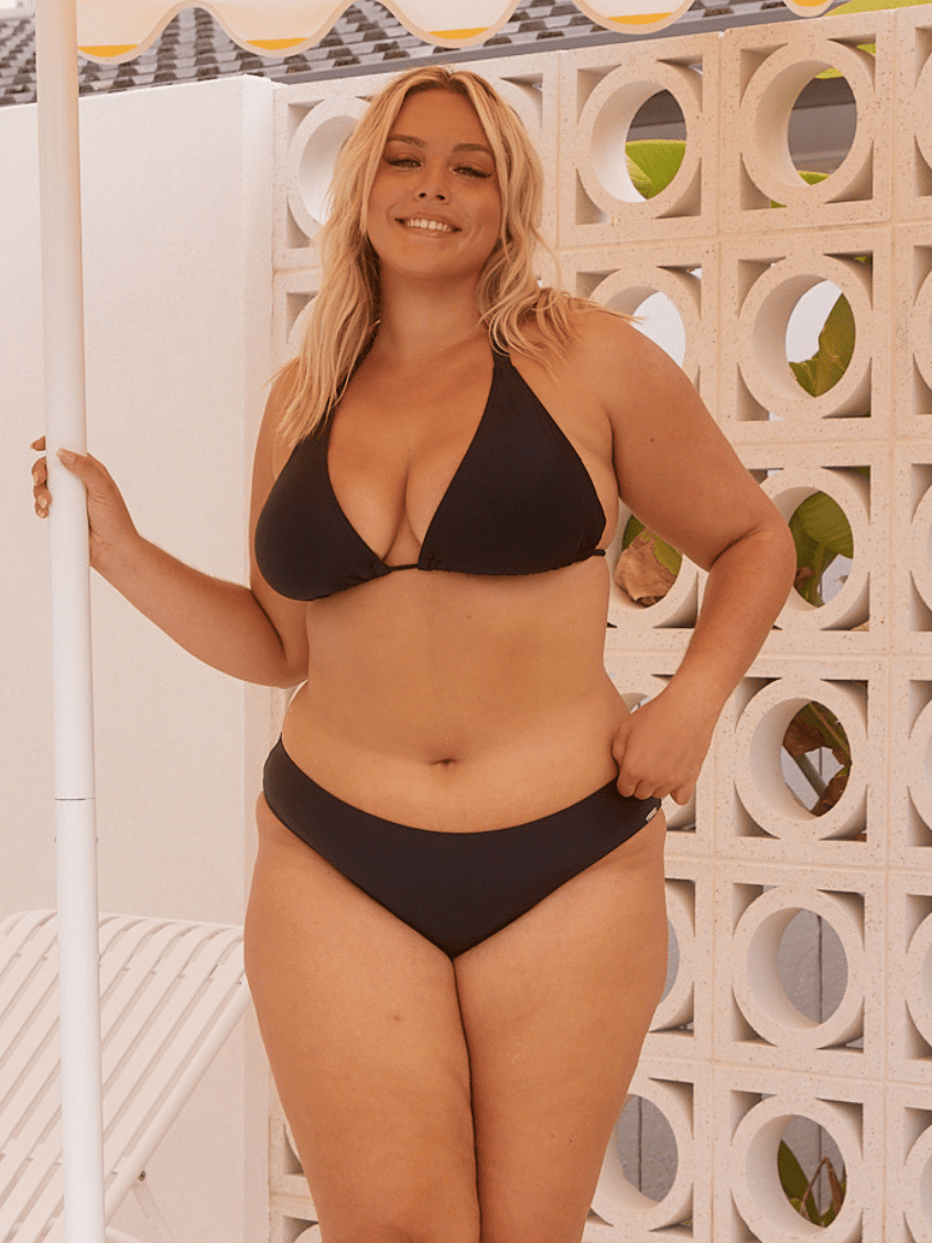 Woman wearing a black bikini with hipster pant standing against a decorative wall.