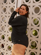 A woman wearing a black long-sleeve swim dress stands in front of a white concrete block wall with circular cutouts.
