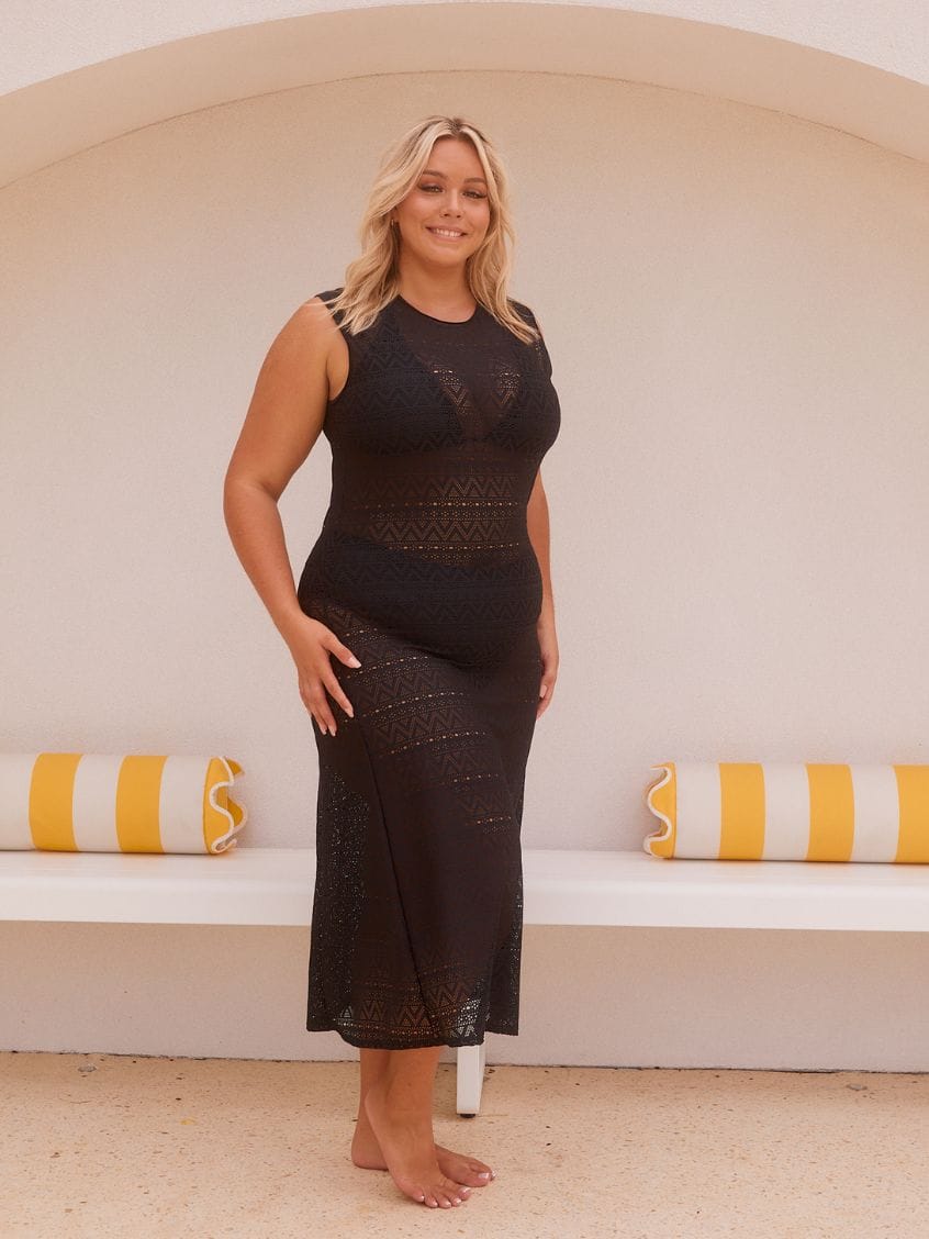 A woman stands barefoot in front of a white arched wall with yellow-and-white striped pillows, wearing a sleeveless black lace maxi dress.