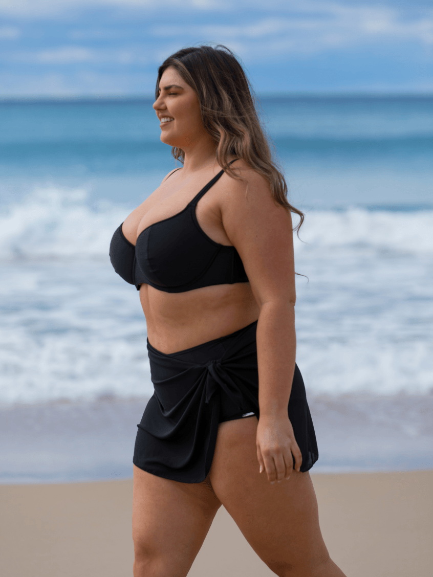 A woman walking on a sandy beach wearing a black two-piece swimwear set consisting of a bandeau-style bikini top and a matching wrap-style bikini skirt.