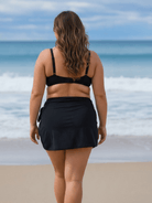A woman stands on a beach facing the ocean, wearing a black swim bikini top and a matching black wrap skirt.