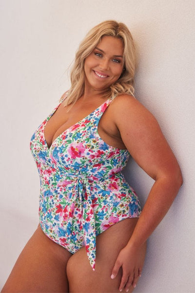 Blonde plus size model wearing low v neck crossover floral one piece with faux tie detail in tones of blue, pink, green and yellow
