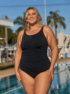 A woman wearing a black one-piece swimsuit stands outdoors by a swimming pool with palm trees and buildings in the background.