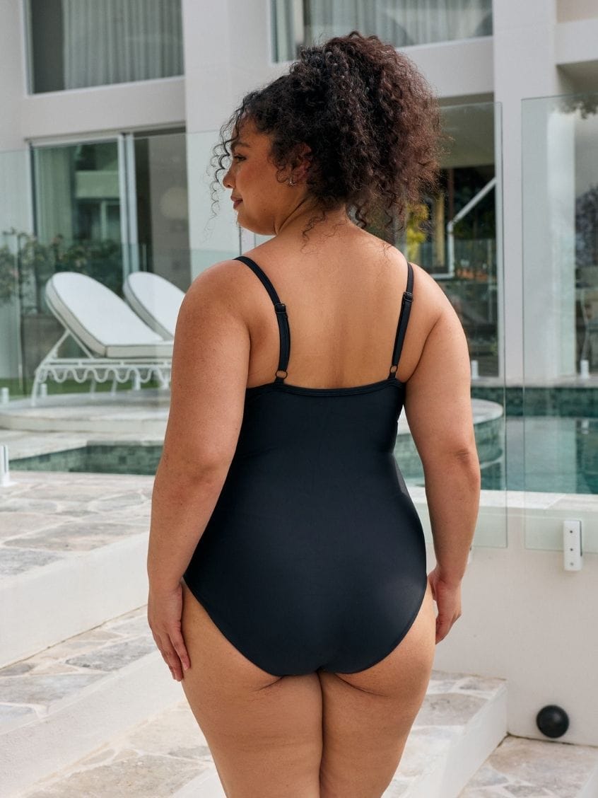 Back of model wearing a classic black one piece with adjustable straps in metallic black with criss cross detail