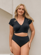 A blonde woman modeling a two-piece black ribbed swimwear set consisting of a short-sleeve wrap-front crop top and matching high-waisted briefs, standing against a textured light beige wall.