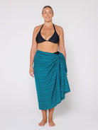 Woman wearing a black bikini top and teal sarong on a white background