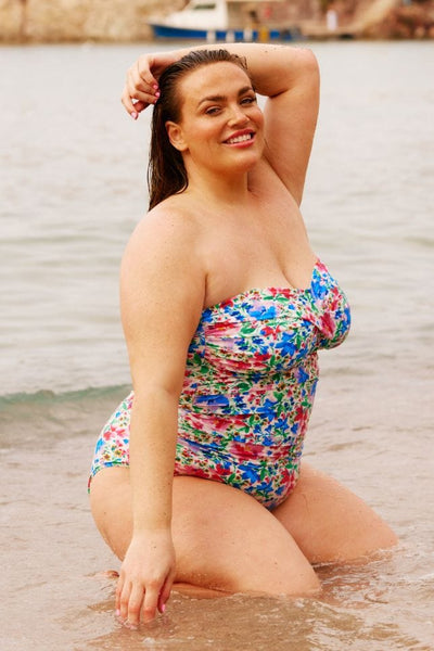 Brunette curve model in water on beach wearing a bandeau swimsuit with removable straps in bright coloured floral print