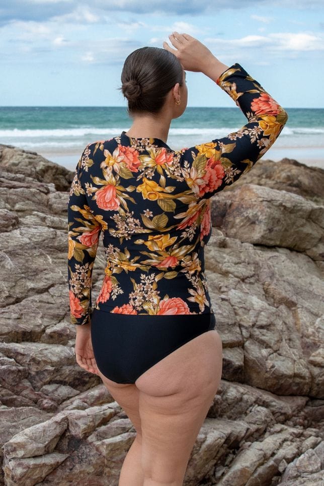 Brunette curve model wearing long sleeve rash vest in floral print with black base and yellow, orange and red tones