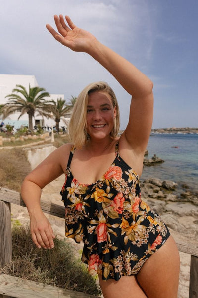 Blonde model at the beach wearing a black based floral swimsuit with orange and yellow flowers with ruffle detail at the front