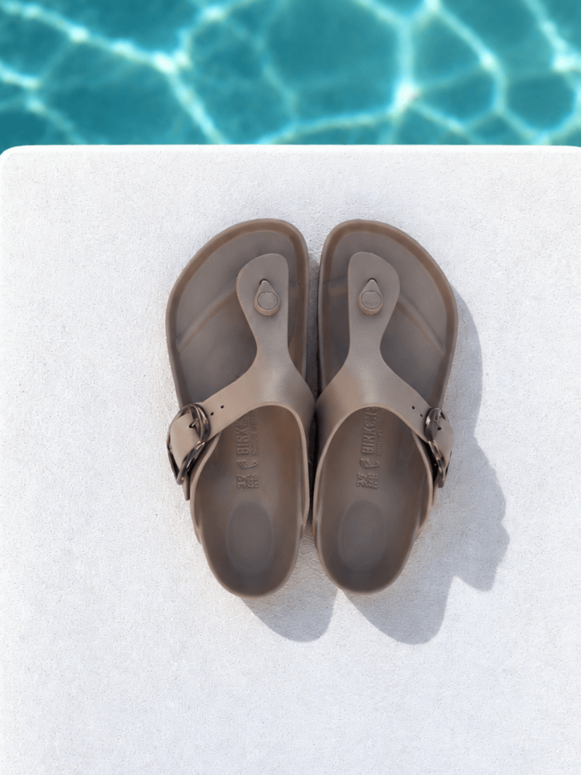 A pair of taupe-brown thong sandals with adjustable straps, laid flat on a white surface with a swimming pool background visible at the top.