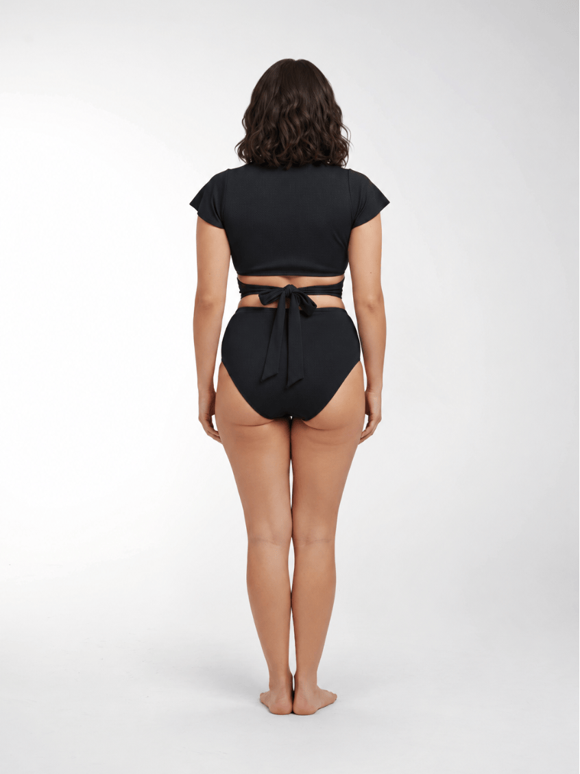 A woman stands with her back to the camera wearing a black two-piece swimsuit with a short-sleeve crop top and high-waisted bottoms featuring a tie-back detail.