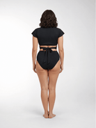 A woman stands with her back to the camera wearing a black two-piece swimsuit with a short-sleeve crop top and high-waisted bottoms featuring a tie-back detail.