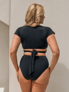 A woman shown from the back wearing a black two-piece swimsuit with a short-sleeve crop top and high-waisted ribbed bottoms tied at the waist, standing against a light beige textured wall.