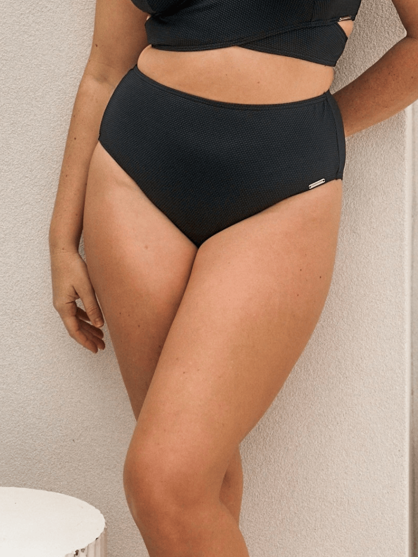 A model wearing a pair of high-waisted textured black bikini bottoms, shown from the waist down against a textured off-white wall.
