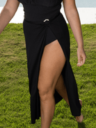 A woman outdoors on green grass wearing a black wrap-style midi skirt with a high leg slit and a small metallic ring detail at the waist.