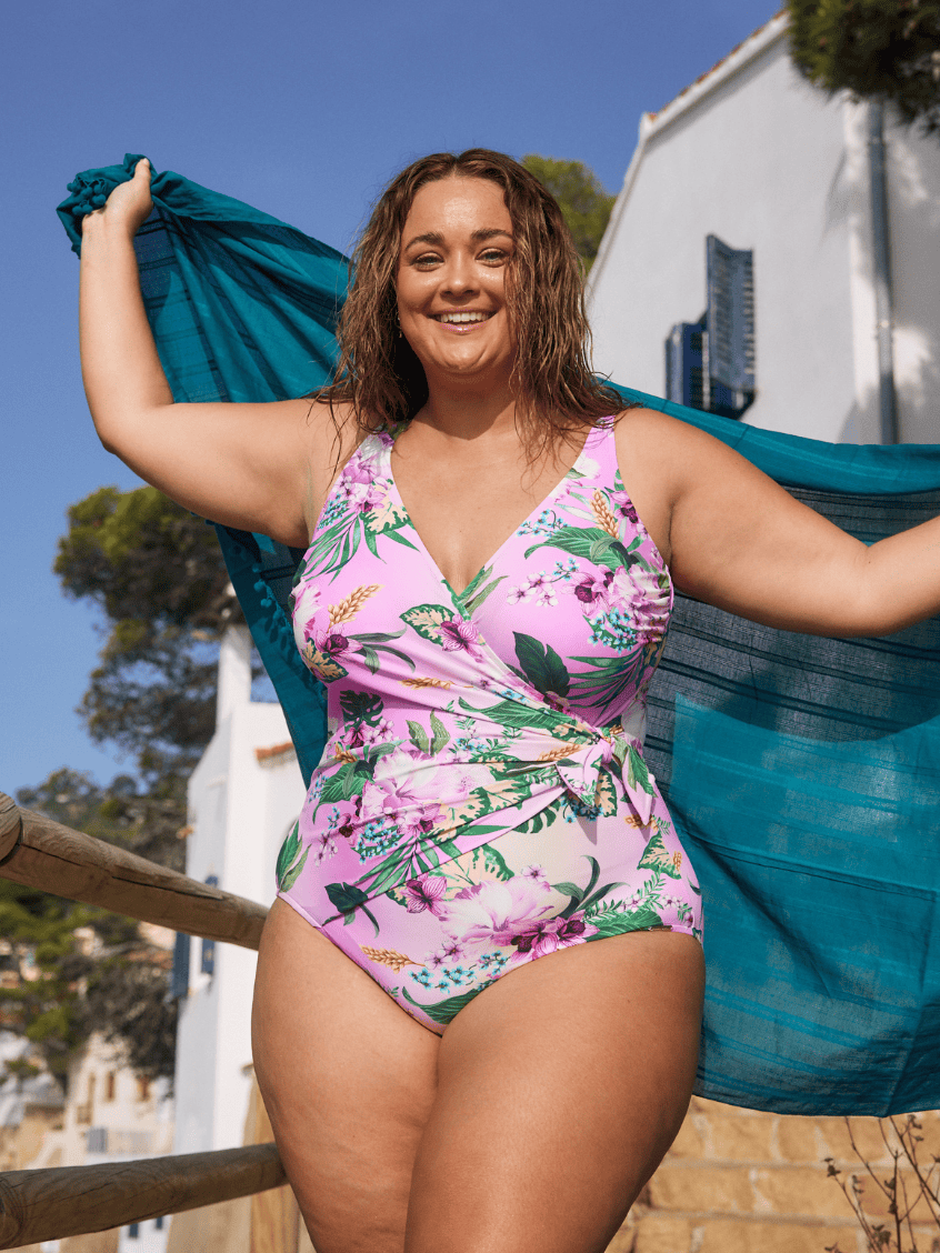 Brunette model by beach wearing one piece swimsuit with tie detail in pink ombre floral colour print