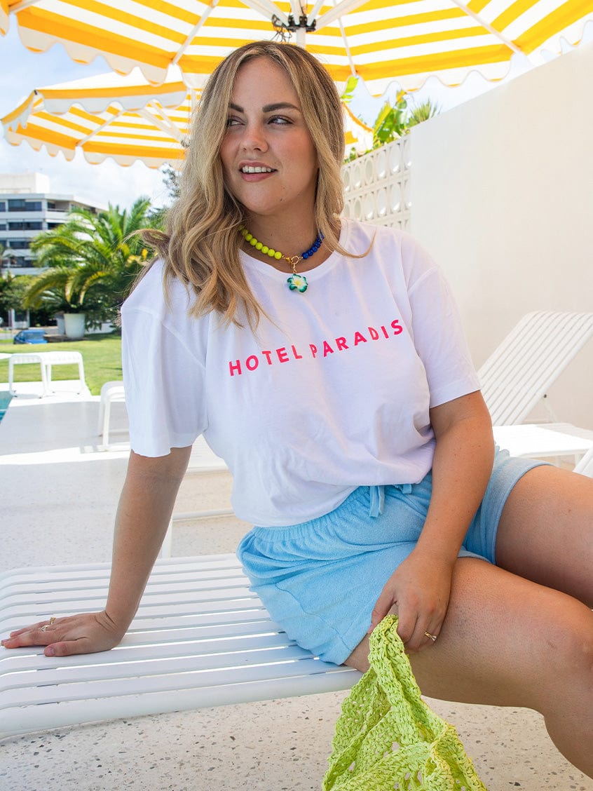 Woman sitting on a lounge chair under a yellow and white umbrella wearing a 'HOTEL PARADISO' t-shirt.