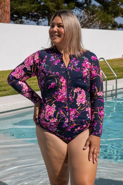Blonde model standing in pool wearing floral long sleeve rashie with zip front in floral pink and purple tones