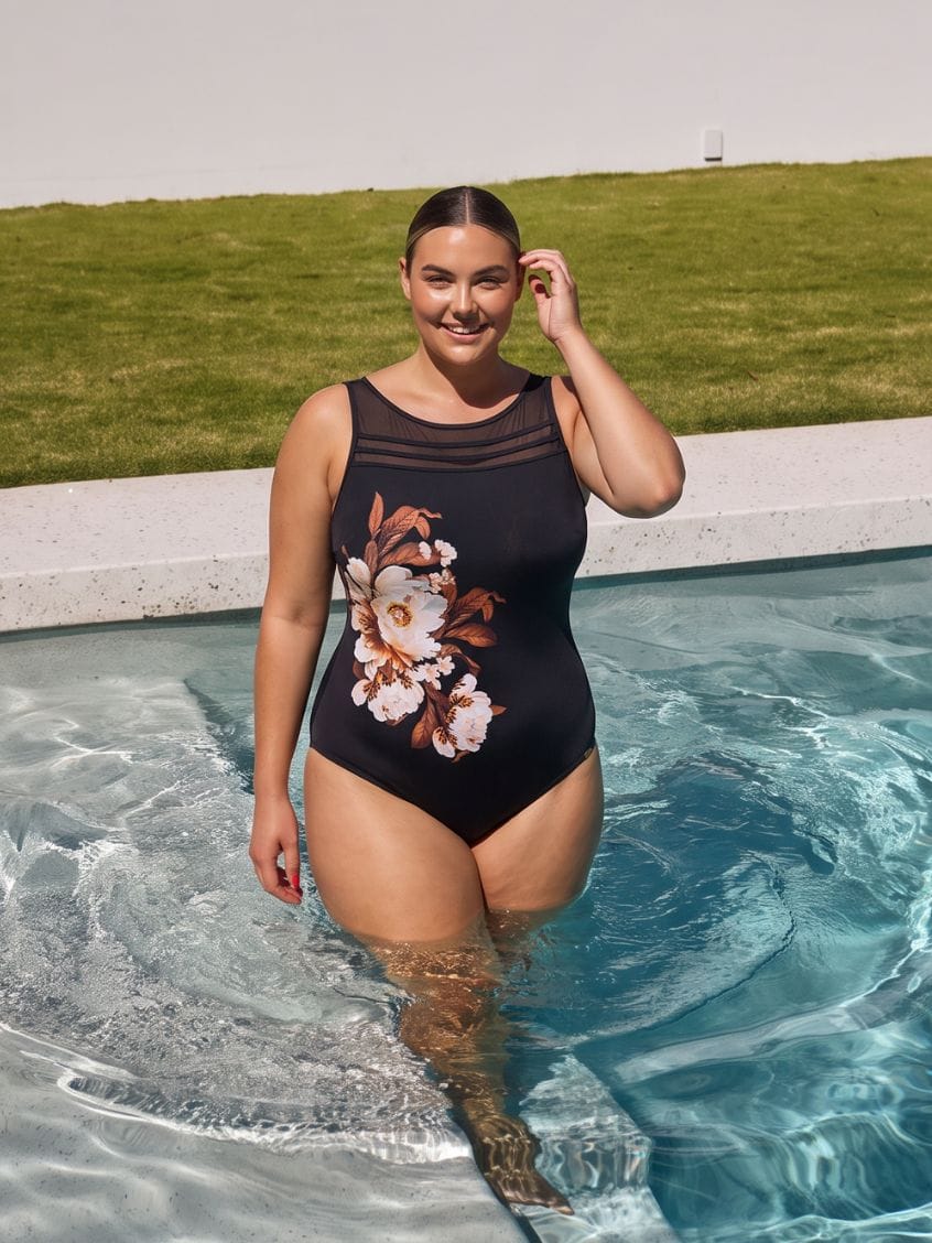A woman stands in a swimming pool wearing a black one-piece swimsuit with a sheer mesh neckline and a large white and brown floral print on the front. A grassy lawn is visible in the background under a bright sky.