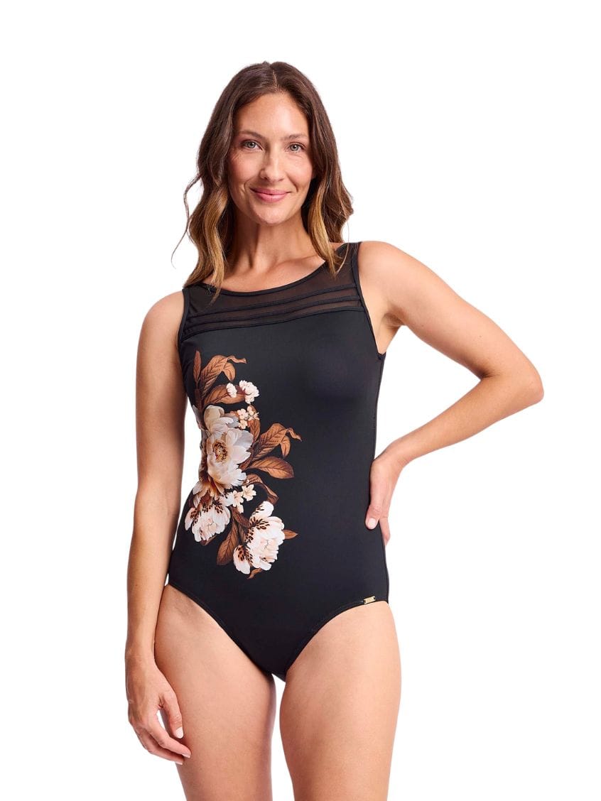 A woman modeling a black one-piece swimsuit with a floral print on the left side, featuring sheer mesh shoulder straps, set against a plain white background.