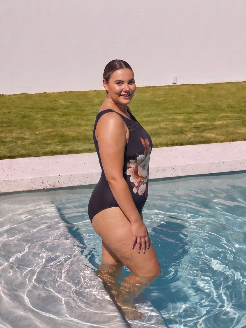 A woman stands in a swimming pool wearing a dark navy one-piece swimsuit with a white and pink floral print on the side, with a green lawn and pale sky in the background.