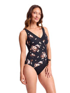 A woman modeling a one-piece swimsuit with a black base and a floral print in pink, beige, and white, set against a plain white background.