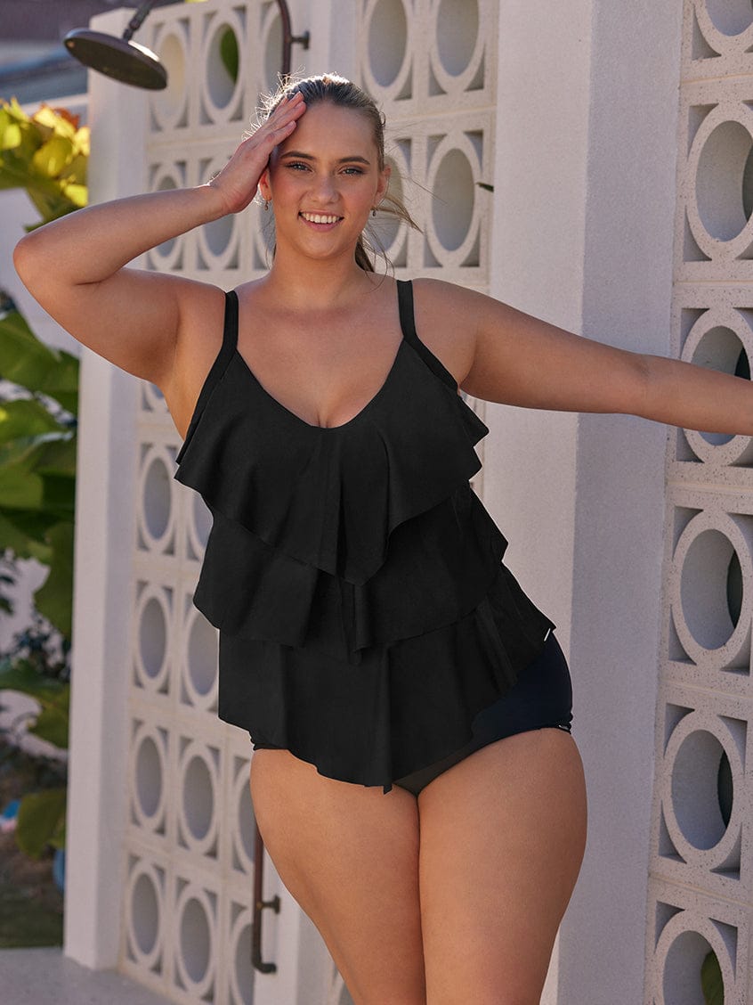 Woman wearing a black ruffled swimsuit standing against a decorative white wall.