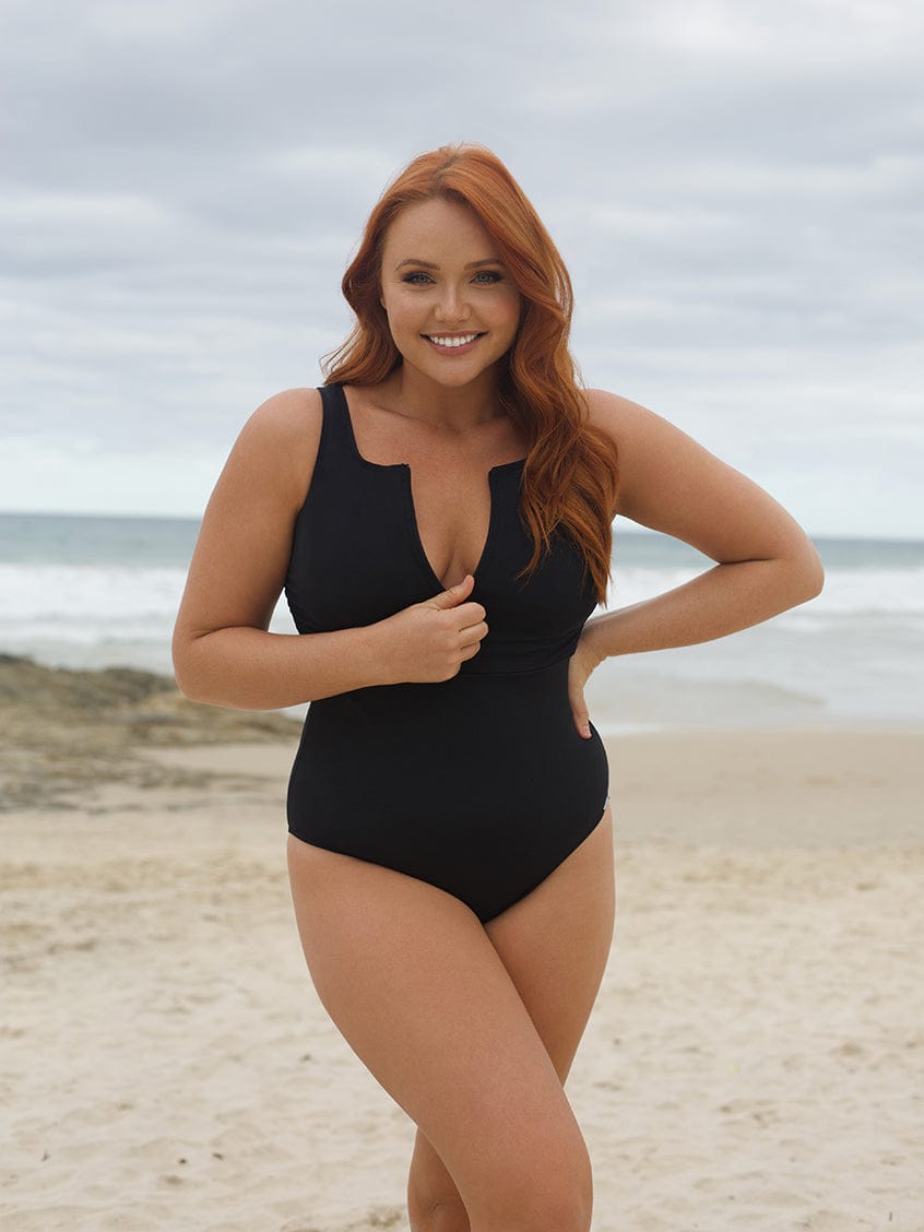 Woman in a black swimsuit standing on a beach with ocean and sky in the background