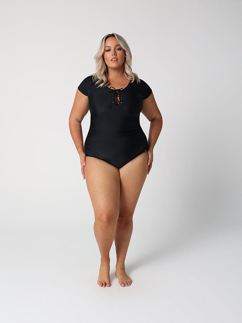 Woman wearing a black swimsuit with a lace-up front, standing in front of a white wall