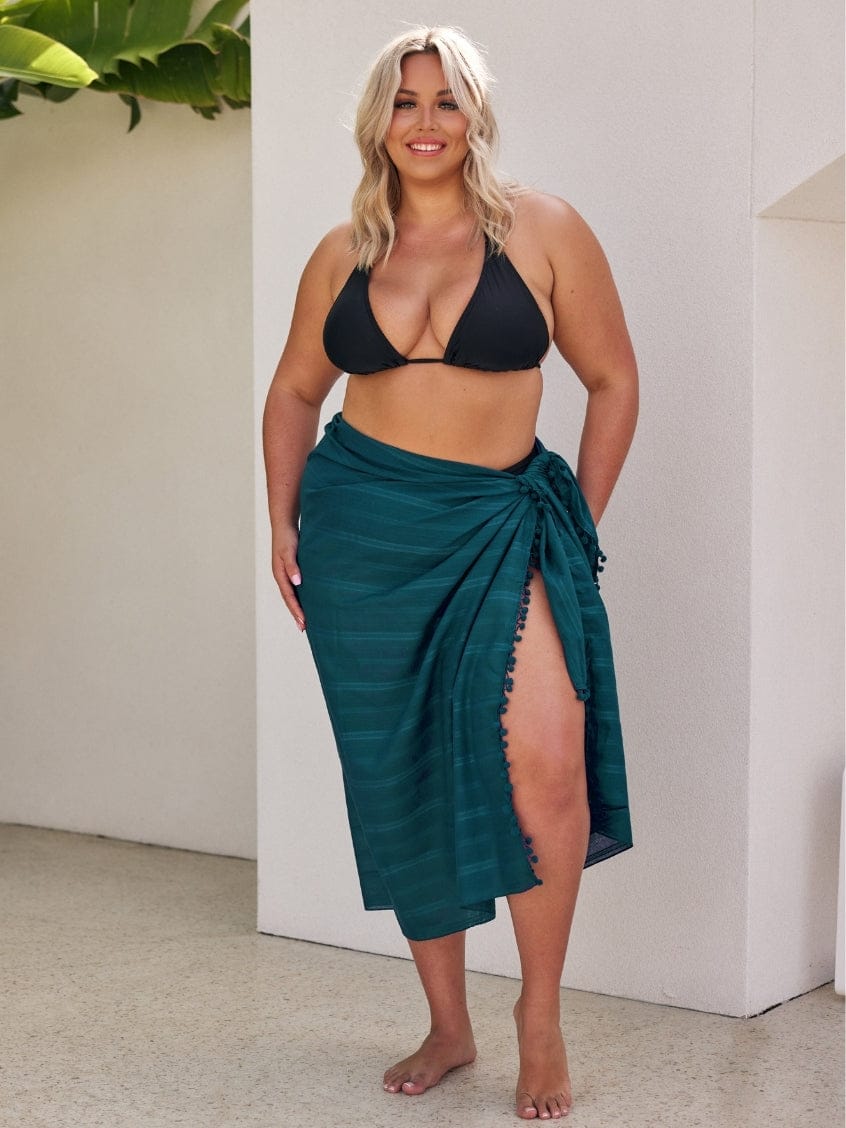 Woman wearing a black bikini top and teal sarong against a white wall.
