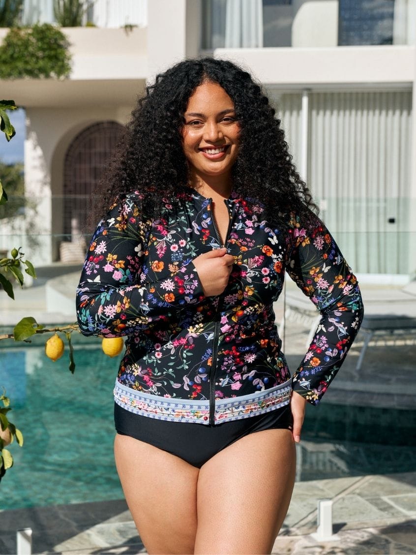 A woman wearing a floral long-sleeve swim jacket over a black bikini stands by a swimming pool in front of a modern white building.