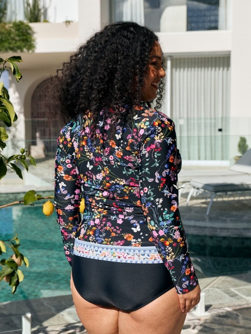A woman standing by a swimming pool wearing a long-sleeve floral swim cover-up with black high-waisted bottoms. The cover-up features a multicolor floral pattern on a black background with a decorative multicolor border at the waist.