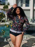 A woman posing by a swimming pool wearing a long-sleeve floral swim cover-up top with black swim briefs.