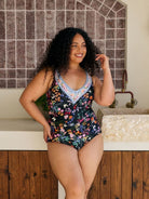 A woman modeling a one-piece swimsuit with a dark navy floral print and a light blue crochet V-neck trim, standing in front of a tiled wall and stone sink.
