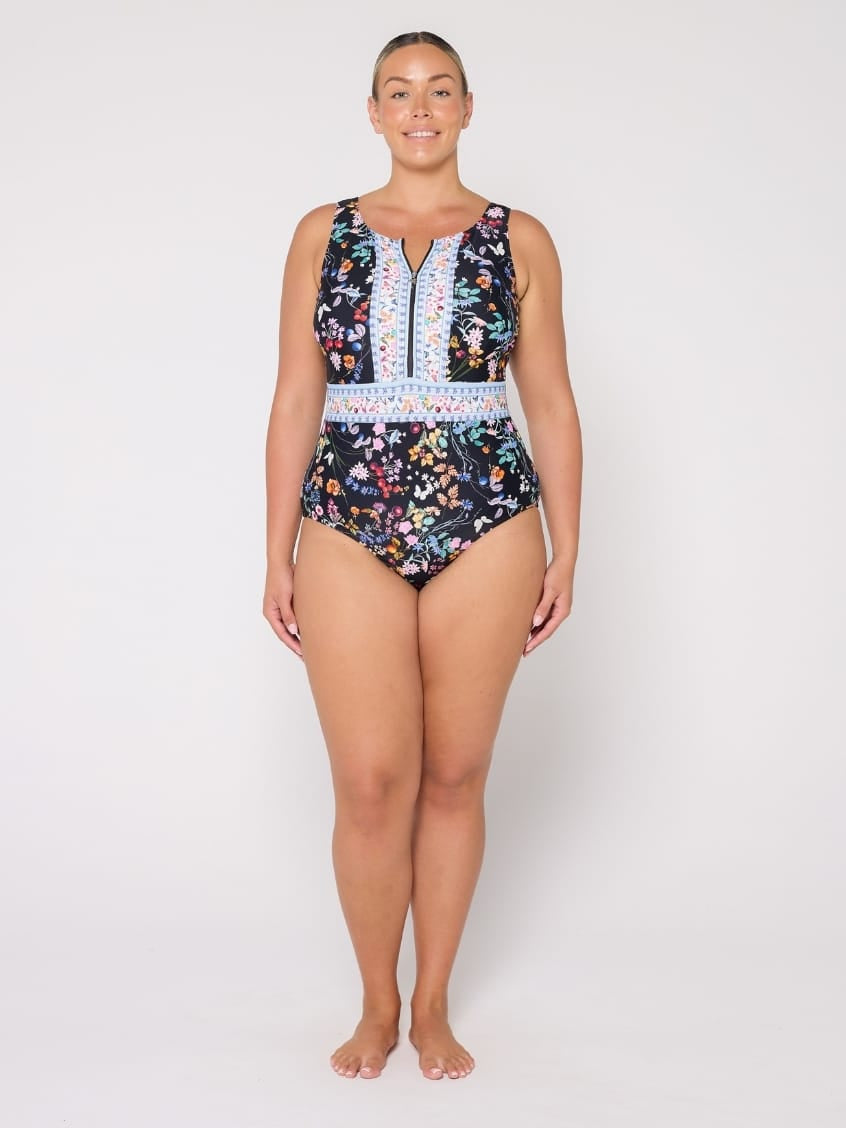 A woman modeling a sleeveless one-piece swimsuit with a dark navy floral print, a white patterned waistband, and a central zipper, photographed against a plain light gray studio background.