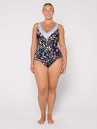 A woman modeling a one-piece swimsuit with a black background and a colorful floral print, featuring a light blue and white patterned V-neckline trim, against a plain light gray studio background.