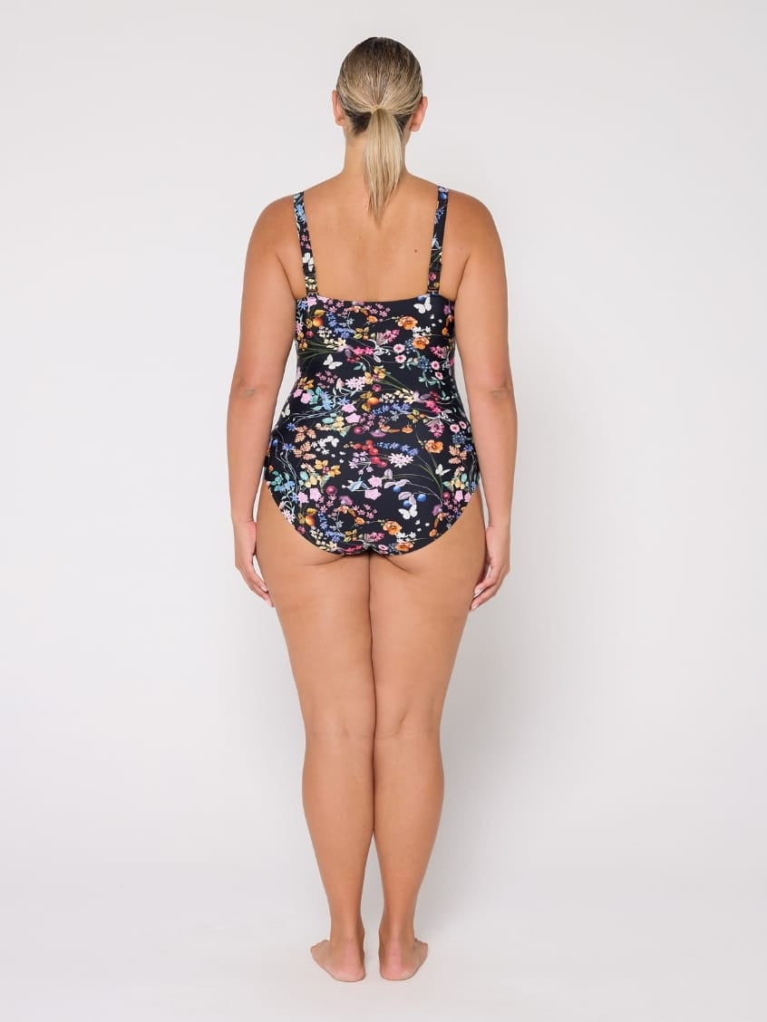 A woman viewed from the back wearing a one-piece swimsuit with a dark floral print, standing against a plain light gray studio background.