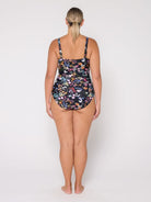 A woman viewed from the back wearing a one-piece swimsuit with a dark floral print, standing against a plain light gray studio background.