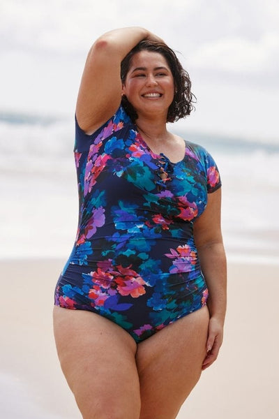 Brunette plus size model wears navy based floral lace up one piece swimsuit on beach