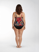 A woman is shown from behind wearing a tankini top swimsuit with a vibrant tropical floral print and solid black bottoms, standing against a light gray studio background.