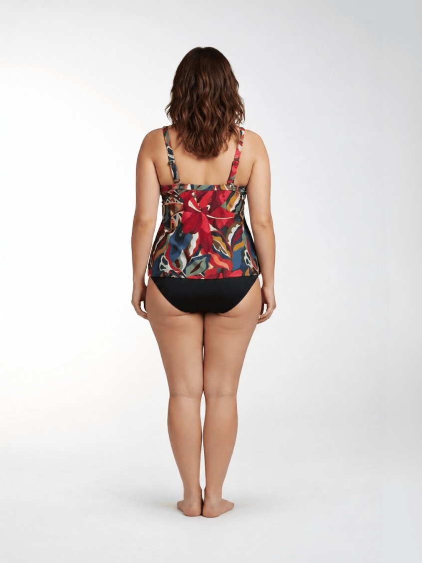 A woman is shown from behind wearing a tankini top swimsuit with a vibrant tropical floral print and solid black bottoms, standing against a light gray studio background.