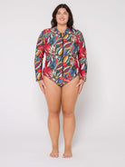 A woman modeling a long-sleeve, collared one-piece swimsuit with a colorful abstract print, standing against a plain light gray studio background.