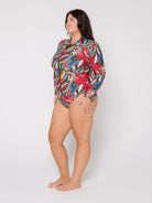 A woman modeling a long-sleeve one-piece swimsuit with a vibrant multicolor tropical floral print, standing against a plain light gray studio background.