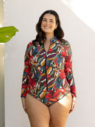 A woman modeling a long-sleeve, one-piece swimsuit with a colorful abstract pattern in red, blue, green, yellow, and orange, standing against a white wall.