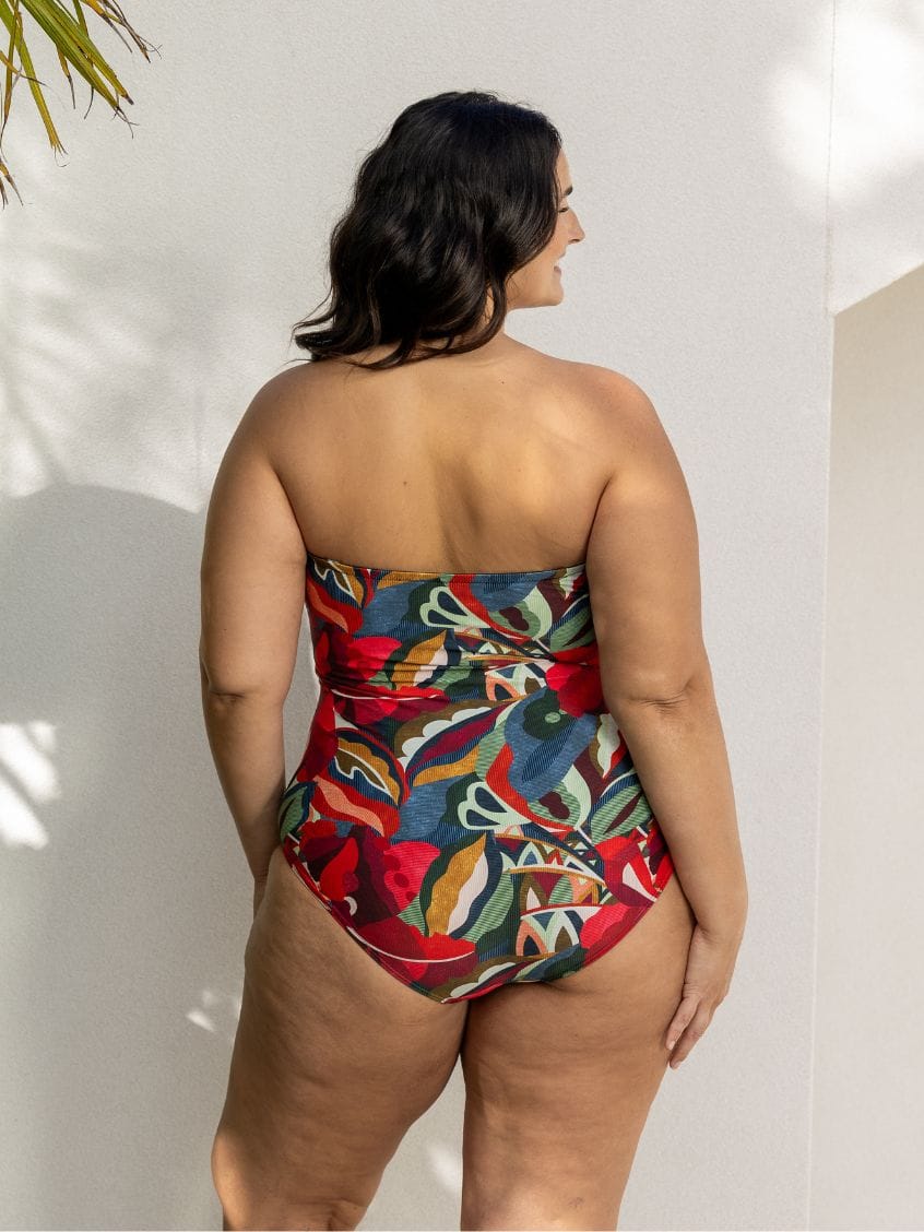A woman is shown from behind wearing a strapless high-waisted one-piece swimsuit with a vibrant, multicolored abstract floral pattern in red, blue, green, and orange, set against a white wall.